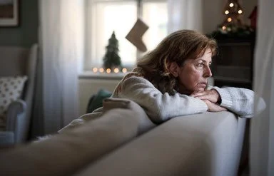 Eine ältere Frau sitzt auf dem Sofa und schaut nachdenklich aus dem Fenster. Im Hintergrund ist Weihnachtsdekoration zu sehen.