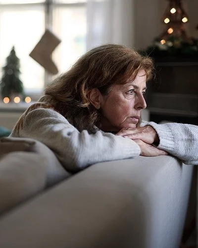 Eine ältere Frau sitzt auf dem Sofa und schaut nachdenklich aus dem Fenster. Im Hintergrund ist Weihnachtsdekoration zu sehen.