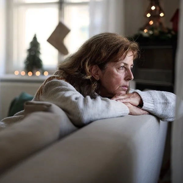 Eine ältere Frau sitzt auf dem Sofa und schaut nachdenklich aus dem Fenster. Im Hintergrund ist Weihnachtsdekoration zu sehen.