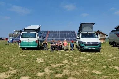 Auf dem Bild sind zwei Autos zu sehen und fünf Personen, die auf Stühlen dazwischen sitzen.