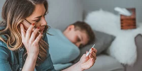 Eine Mutter sitzt bei ihrem zugedeckten Kind auf dem Sofa, hält ein Fieberthermometer in der Hand und telefoniert.