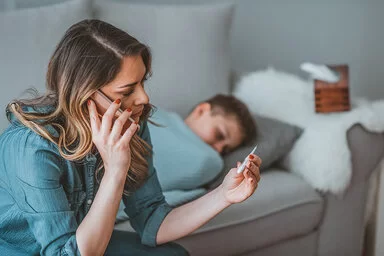Eine Mutter sitzt bei ihrem zugedeckten Kind auf dem Sofa, hält ein Fieberthermometer in der Hand und telefoniert.