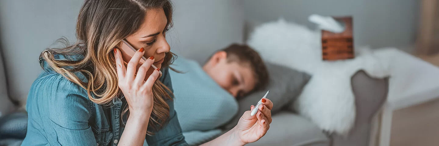 Eine Mutter sitzt bei ihrem zugedeckten Kind auf dem Sofa, hält ein Fieberthermometer in der Hand und telefoniert.
