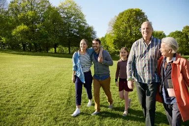 Eine Mehrgenerationenfamilie läuft gemeinsam und fröhlich lachend durch einen sonnigen Park. Im Bild sind zwei ältere Erwachsene, ein junges Paar und ein Kind zu sehen. Im Hintergrund stehen grüne Bäume, der Himmel ist klar und blau.