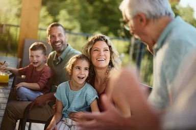 Eine Mehrgenerationenfamilie sitzt fröhlich lachend auf einer Terasse. Im Bild sind ein älterer Mann, ein Paar mittleren Alters und zwei Kinder zu sehen. Im Hintergrund sind grüne Büsche und Bäume zu erkennen.