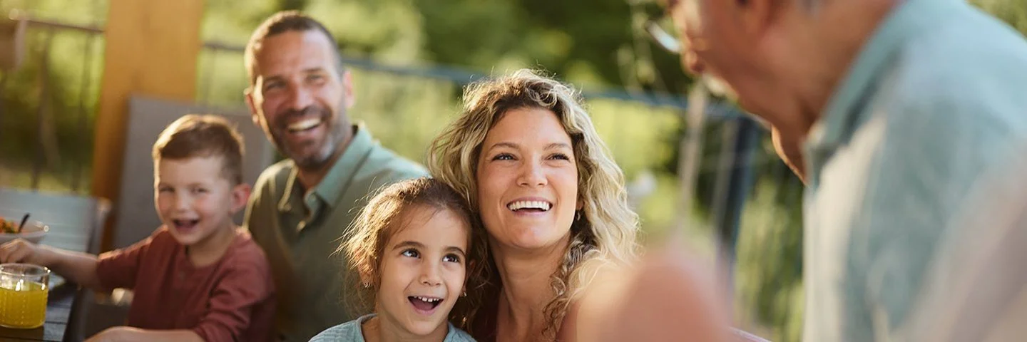 Eine Mehrgenerationenfamilie sitzt fröhlich lachend auf einer Terasse. Im Bild sind ein älterer Mann, ein Paar mittleren Alters und zwei Kinder zu sehen. Im Hintergrund sind grüne Büsche und Bäume zu erkennen.