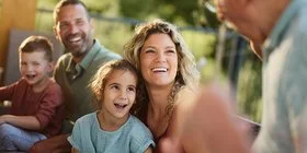 Eine Mehrgenerationenfamilie sitzt fröhlich lachend auf einer Terasse. Im Bild sind ein älterer Mann, ein Paar mittleren Alters und zwei Kinder zu sehen. Im Hintergrund sind grüne Büsche und Bäume zu erkennen.