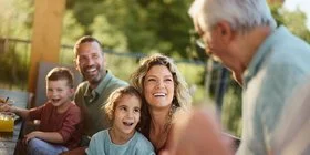 Eine Mehrgenerationenfamilie sitzt fröhlich lachend auf einer Terasse. Im Bild sind ein älterer Mann, ein Paar mittleren Alters und zwei Kinder zu sehen. Im Hintergrund sind grüne Büsche und Bäume zu erkennen.