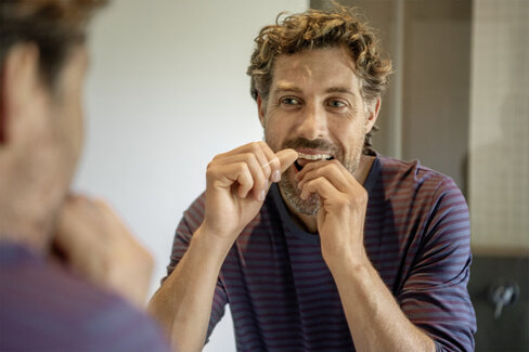 Ein Mann steht vor dem Spiegel und behandelt seine Zähne mit Zahnseide. Die AOK-Zahnklinik ist eine Prophylaxepraxis.