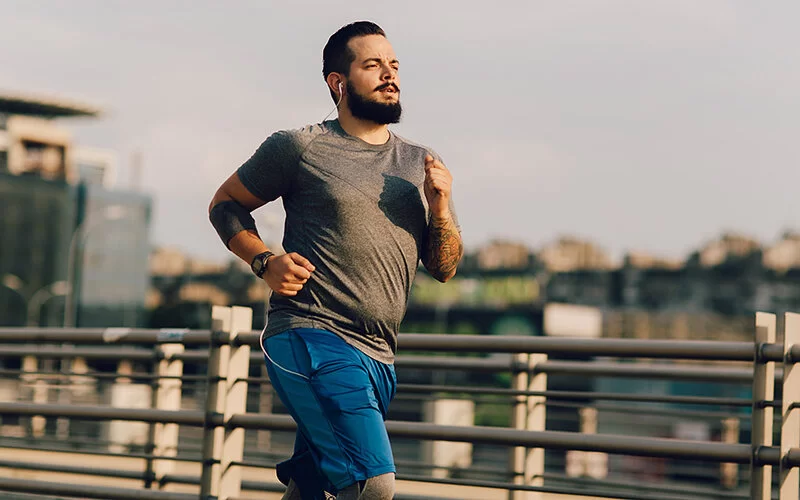 Ein mehrgewichtiger junger Mann joggt morgens über eine Brücke in einer Großstadt, währenddessen hört er Musik.