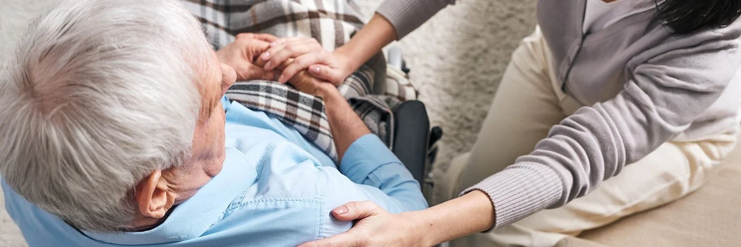 Hilfsbereite Angehörige sitzt neben einem älteren Mann im Rollstuhl und legt ihm die Hand auf die Schulter.