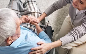 Hilfsbereite Angehörige sitzt neben einem älteren Mann im Rollstuhl und legt ihm die Hand auf die Schulter.