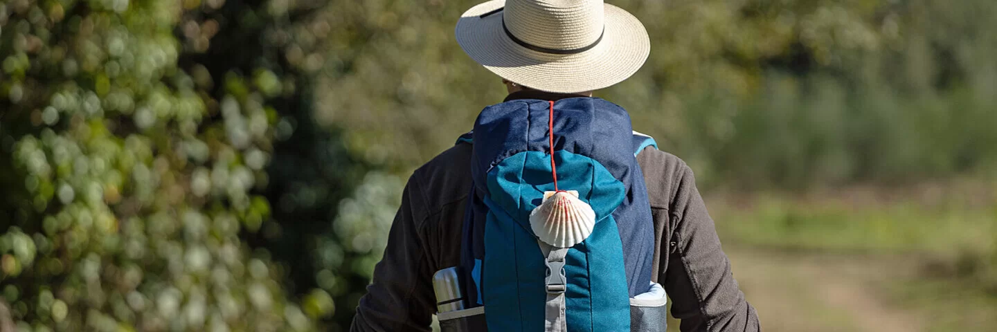 Rückansicht eines Pilgers mit Jakobsmuschel am Rucksack, der einen Feldweg nach Santiago de Compostela nimmt.