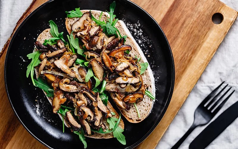 Zwei Scheiben Brot auf einem schwarzen Teller, belegt mit gebratenen Pilzen und frischem Rucola.