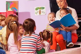 Eine Frau sitzt mit neun Kindern in einem rosafarbenen Raum und liest ihnen aus einem blauen Buch vor.