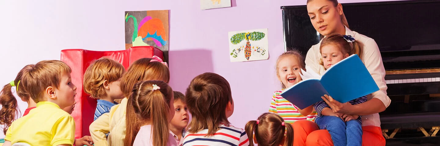 Eine Frau sitzt mit neun Kindern in einem rosafarbenen Raum und liest ihnen aus einem blauen Buch vor.