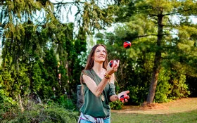 Frau steht in der Natur vor ein paar Bäumen und wirft Jonglierbälle in die Luft.