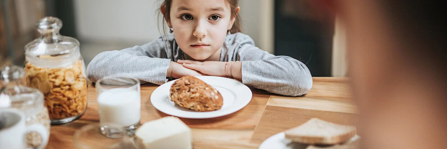 Ein Kind sitzt am Frühstückstisch und möchte das Brötchen vor sich nicht essen.