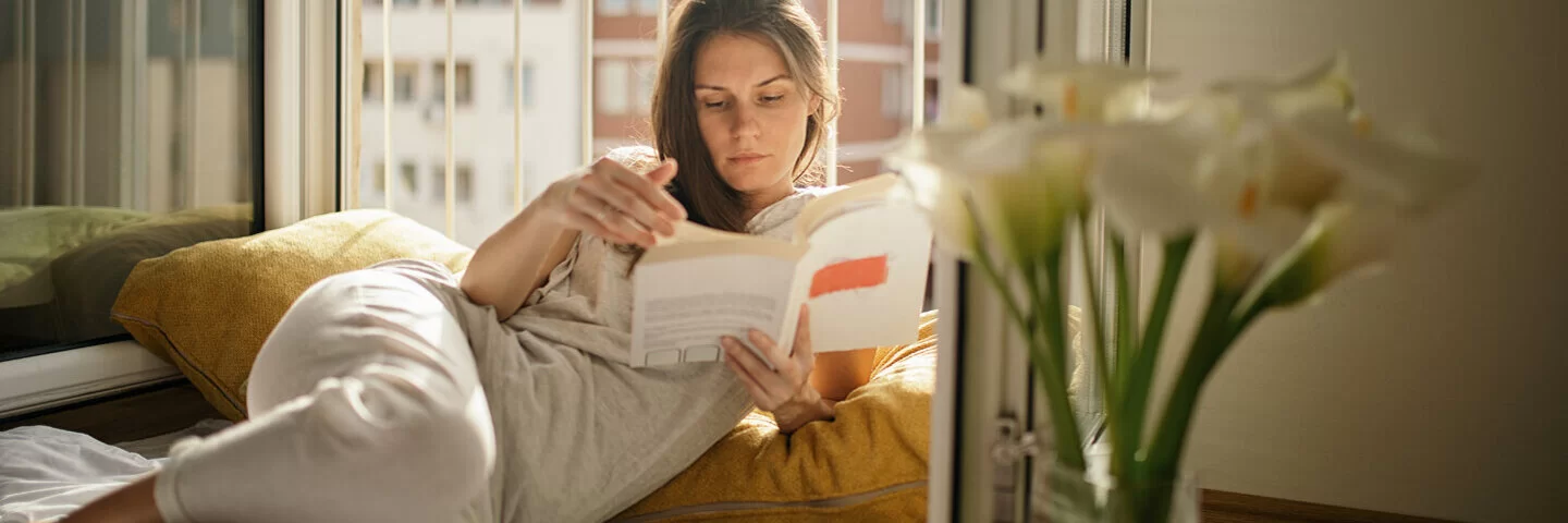 Eine Frau liegt gemütlich neben dem offenen Fenster und liest ein Buch, da Lesen gut für die Gesundheit und Entspannung ist.