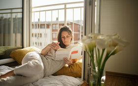 Eine Frau liegt gemütlich neben dem offenen Fenster und liest ein Buch, da Lesen gut für die Gesundheit und Entspannung ist.