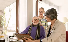 Ein altes Paar spricht mit einer Pflegeberaterin der AOK. Die Pflegeberaterin erklärt in einem Beratungsgespräch, wie sich die Pflege organisieren lässt.