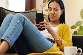 Eine junge Frau sitzt auf einem Sofa und schaut auf ihr Smartphone.