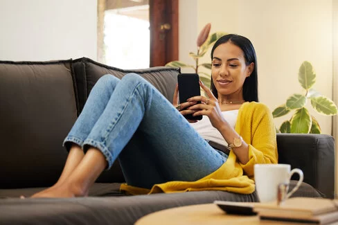 Eine junge Frau sitzt auf einem Sofa und schaut auf ihr Smartphone.