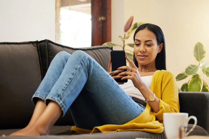 Eine junge Frau sitzt auf einem Sofa und schaut auf ihr Smartphone.