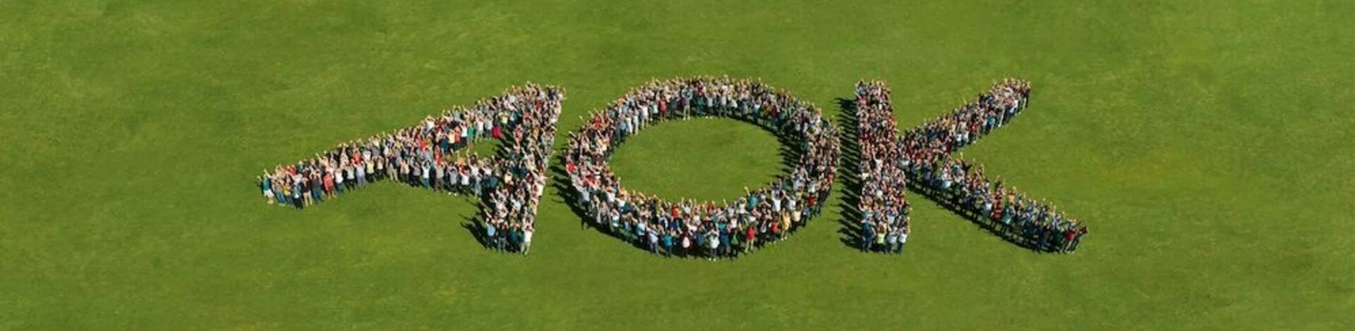 Hunderte Menschen bilden die Buchstaben A, O, K auf einer Rasenfläche. Aufnahme aus der Vogelperspektive.