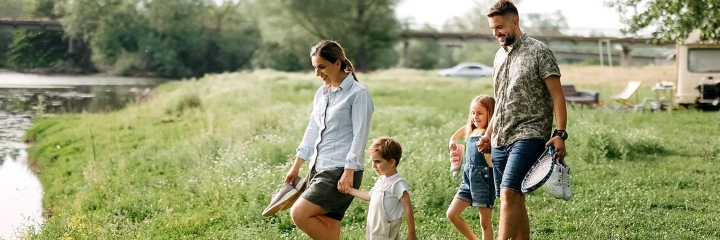 Eine Familie mit zwei Kindern geht barfuß über eine Wiese am Ufer eines Sees; die Familie trägt Freizeitausrüstung und Schuhe in den Händen. 