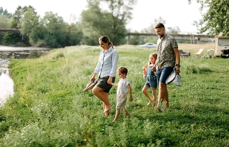 Eine Familie mit zwei Kindern geht barfuß über eine Wiese am Ufer eines Sees; die Familie trägt Freizeitausrüstung und Schuhe in den Händen. 
