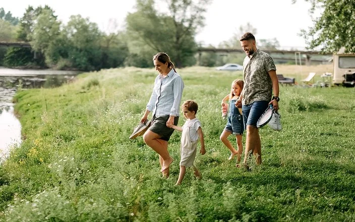 Eine Familie mit zwei Kindern geht barfuß über eine Wiese am Ufer eines Sees; die Familie trägt Freizeitausrüstung und Schuhe in den Händen.