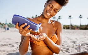 Eine junge Frau am Strand drückt Sonnencreme aus einer Flasche in die Hand, um sich vor der Sonne zu schützen.