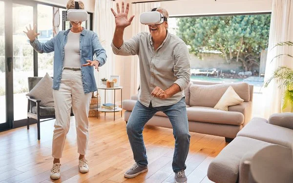 Zwei ältere Menschen mit einer VR-Brille bewegen sich im Wohnzimmer und lächeln.
