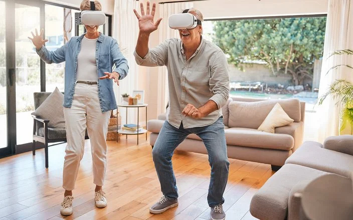 Zwei ältere Menschen mit einer VR-Brille bewegen sich im Wohnzimmer und lächeln.