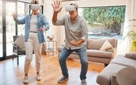 Zwei ältere Menschen mit einer VR-Brille bewegen sich im Wohnzimmer und lächeln.