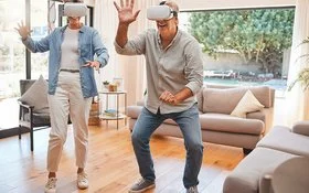 Zwei ältere Menschen mit einer VR-Brille bewegen sich im Wohnzimmer und lächeln.