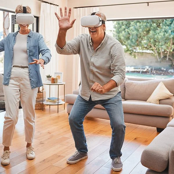 Zwei ältere Menschen mit einer VR-Brille bewegen sich im Wohnzimmer und lächeln.