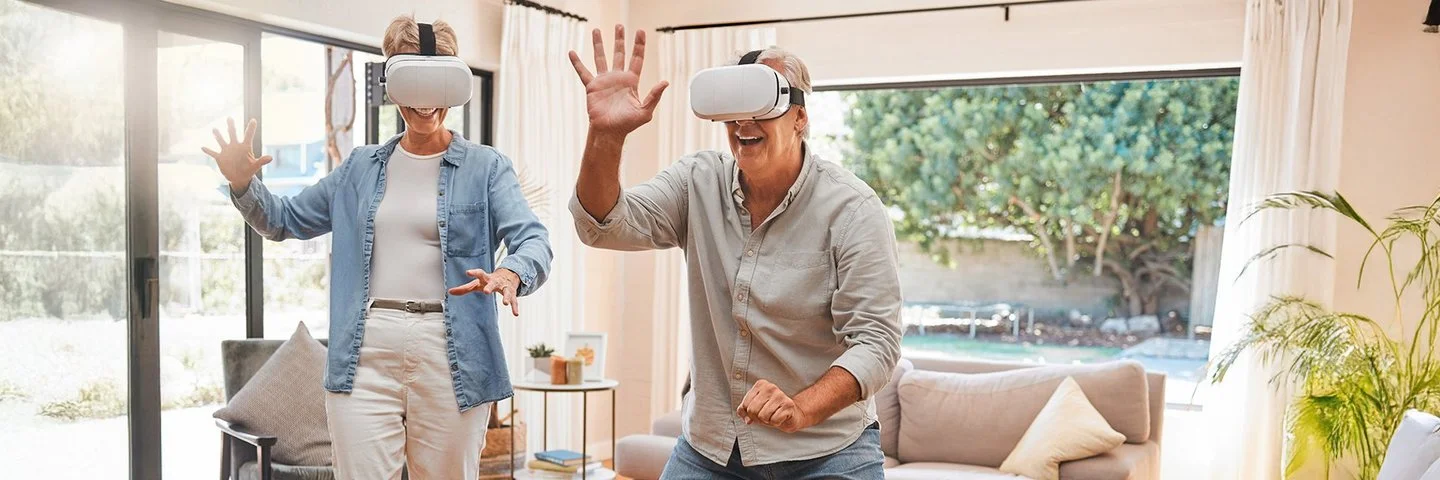 Zwei ältere Menschen mit einer VR-Brille bewegen sich im Wohnzimmer und lächeln.