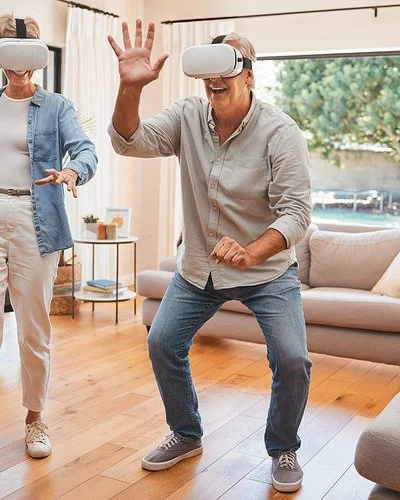 Zwei ältere Menschen mit einer VR-Brille bewegen sich im Wohnzimmer und lächeln.