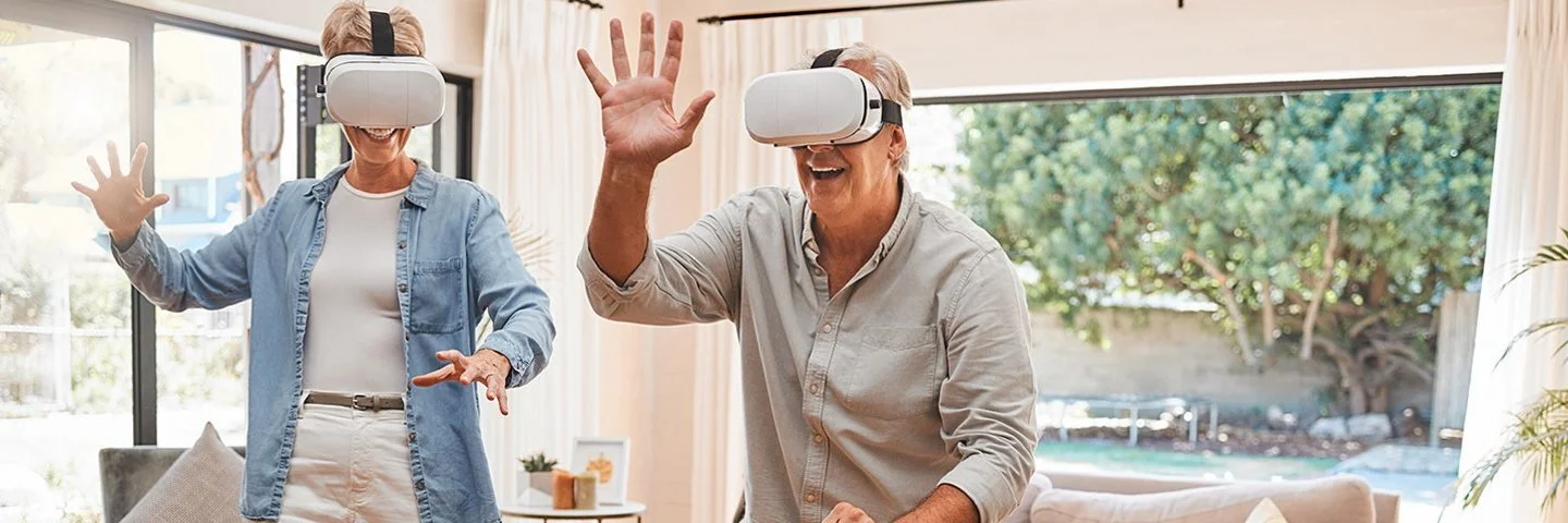 Zwei ältere Menschen mit einer VR-Brille bewegen sich im Wohnzimmer und lächeln.