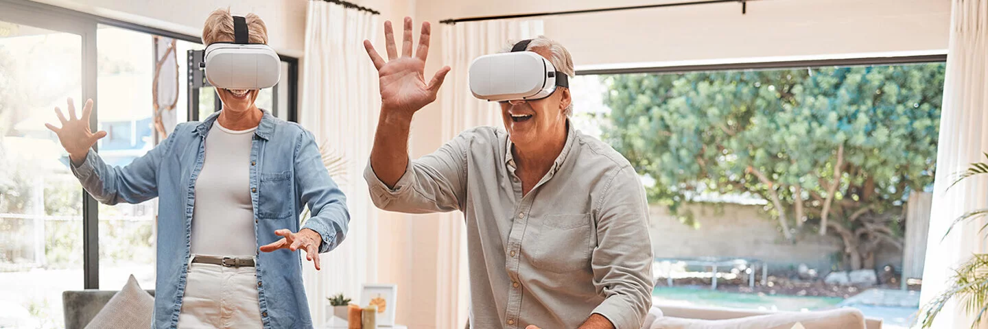 Zwei ältere Menschen mit einer VR-Brille bewegen sich im Wohnzimmer und lächeln.