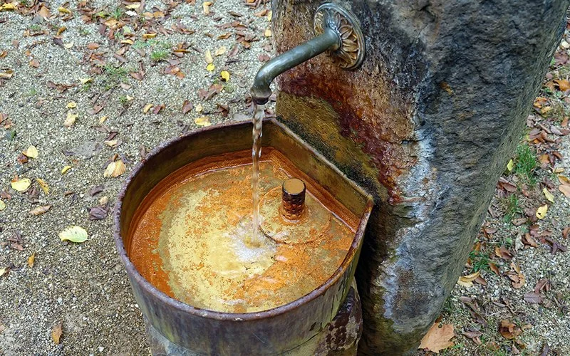 Metallener Quellhahn speist eine kleine Schale; orange-braune Mineralablagerungen am Stein und Becken.