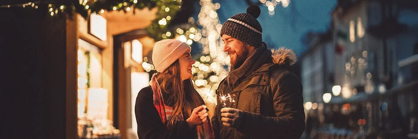 Paar mit Mützen hält Wunderkerzen auf einem abendlichen, festlich beleuchteten Weihnachtsmarkt in Hessen.