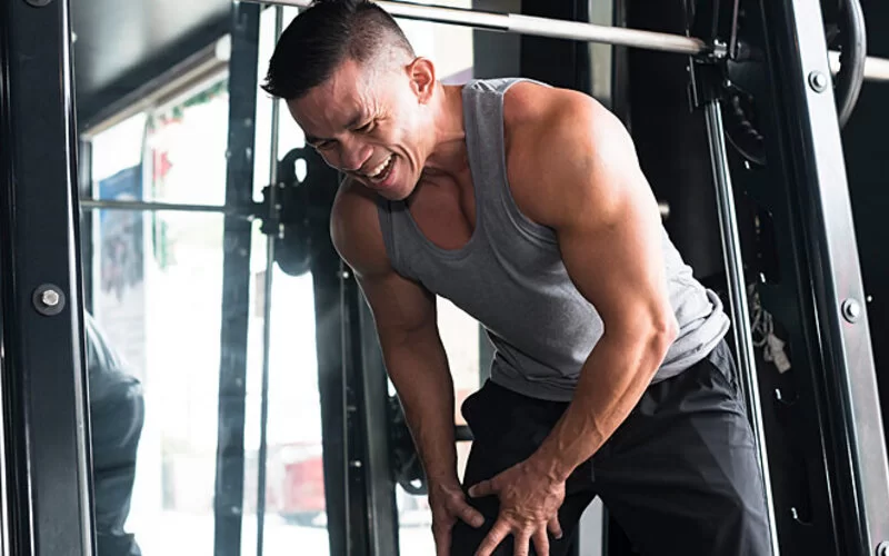 Ein Mann verzieht im Fitnessstudio vor Schmerz das Gesicht und fasst sich mit beiden Händen an den rechten Oberschenkel.