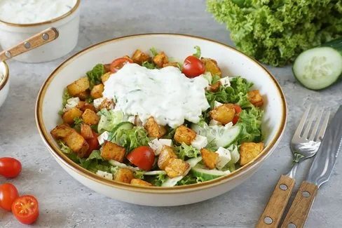 Ein Teller mit gerösteten Kartoffelecken auf gemischtem Salat, garniert mit Kirschtomaten und Tzatziki.