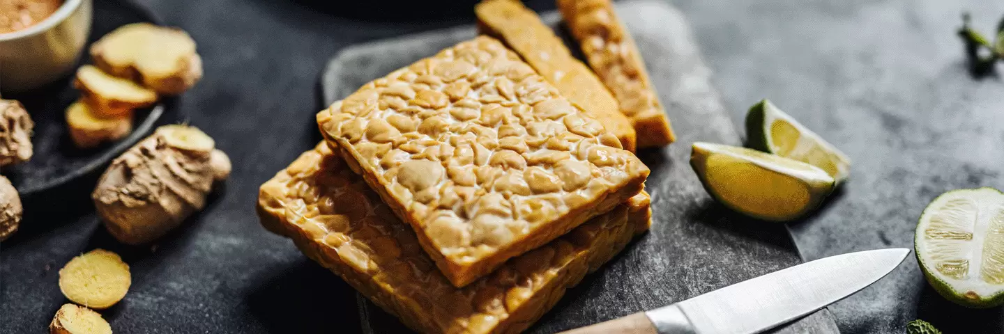 In Scheiben geschnittener Tempeh auf einem dunklen Schneidebrett, daneben aufgeschittener Ingwer und Limette