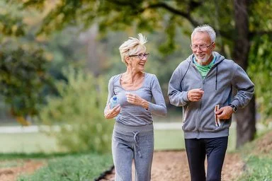 Ein älteres Paar geht gemeinsam im Park joggen. Sie unterhalten sich während des Laufens und lachen dabei. Im Hintergrund ist eine Parkanlage mit Bäumen, Büschen und Wiesen zu sehen.