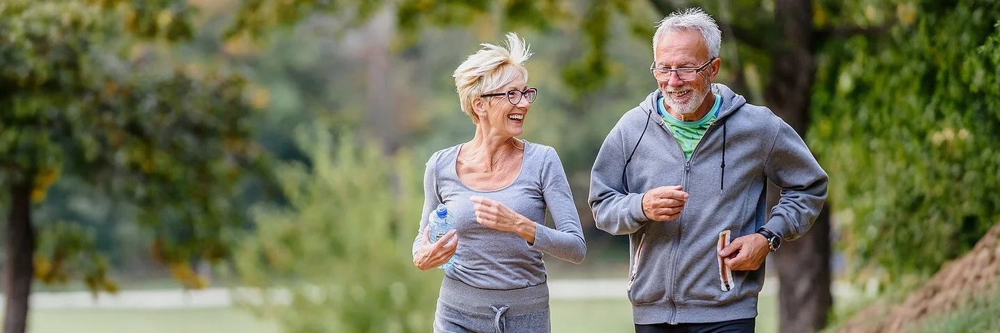 Ein älteres Paar geht gemeinsam im Park joggen. Sie unterhalten sich während des Laufens und lachen dabei. Im Hintergrund ist eine Parkanlage mit Bäumen, Büschen und Wiesen zu sehen.