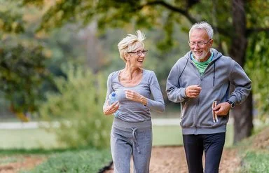Ein älteres Paar geht gemeinsam im Park joggen. Sie unterhalten sich während des Laufens und lachen dabei. Im Hintergrund ist eine Parkanlage mit Bäumen, Büschen und Wiesen zu sehen.